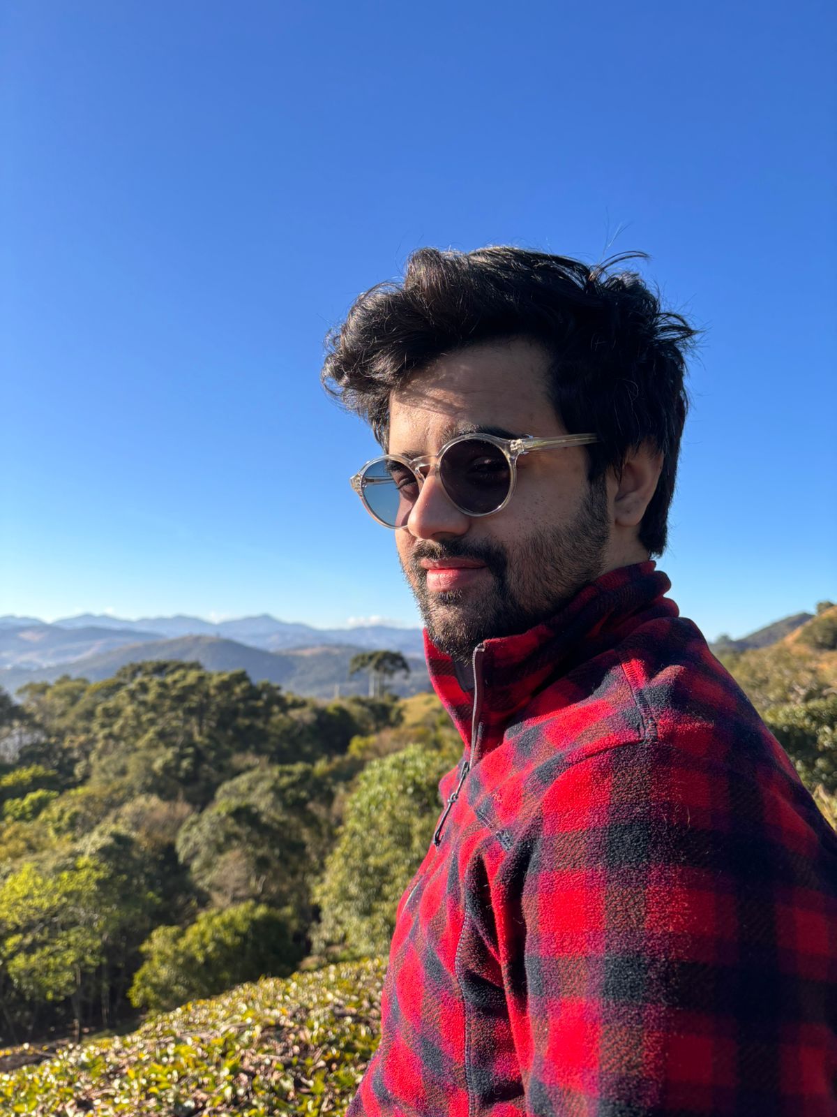 Thainan Prado ao ar livre, de camisa xadrez vermelha, com montanhas ao fundo